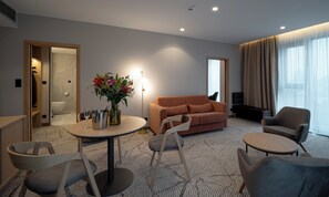 Moderni hotellihuone, jossa on sohva, nojatuoleja, ruokapöytä tuoleineen, maljakko kukkineen ja näkymä ulos.