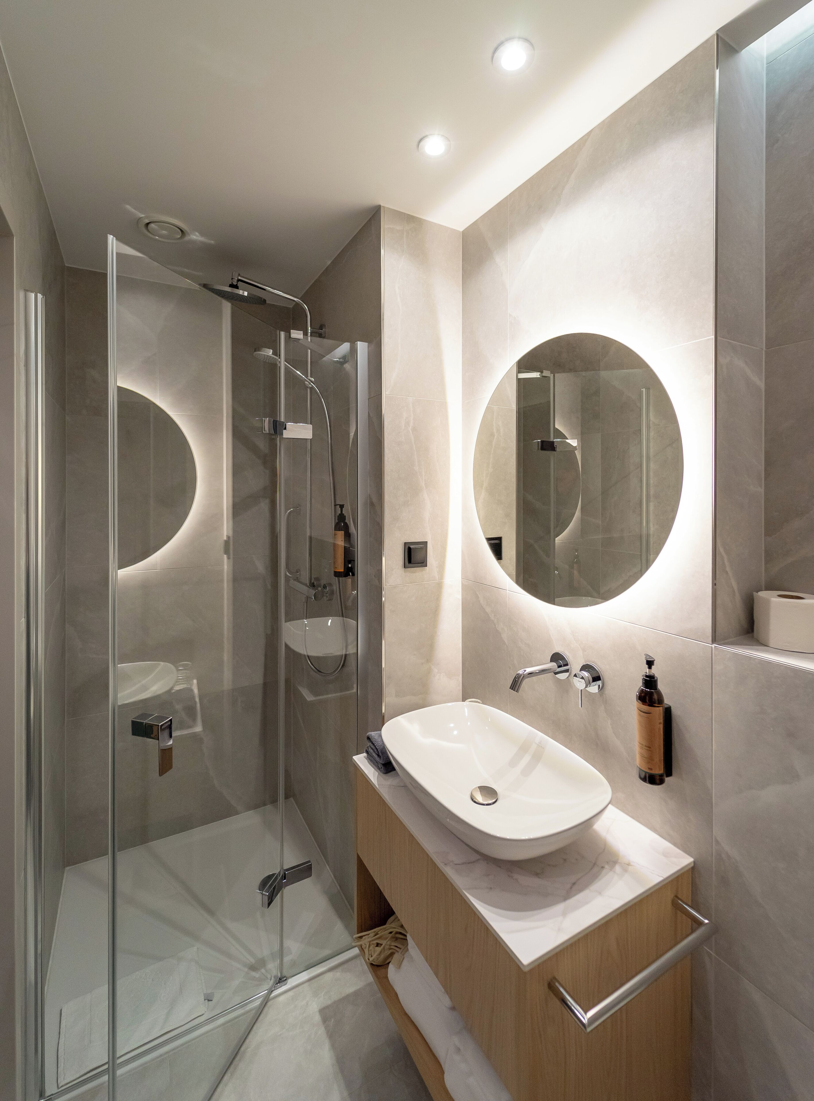 A modern bathroom with a glass-enclosed shower, a round mirror, and a wooden vanity with a white sink.
