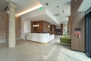 Zona de recepción de hotel con mostrador de mármol, detalles de madera y un sillón verde.