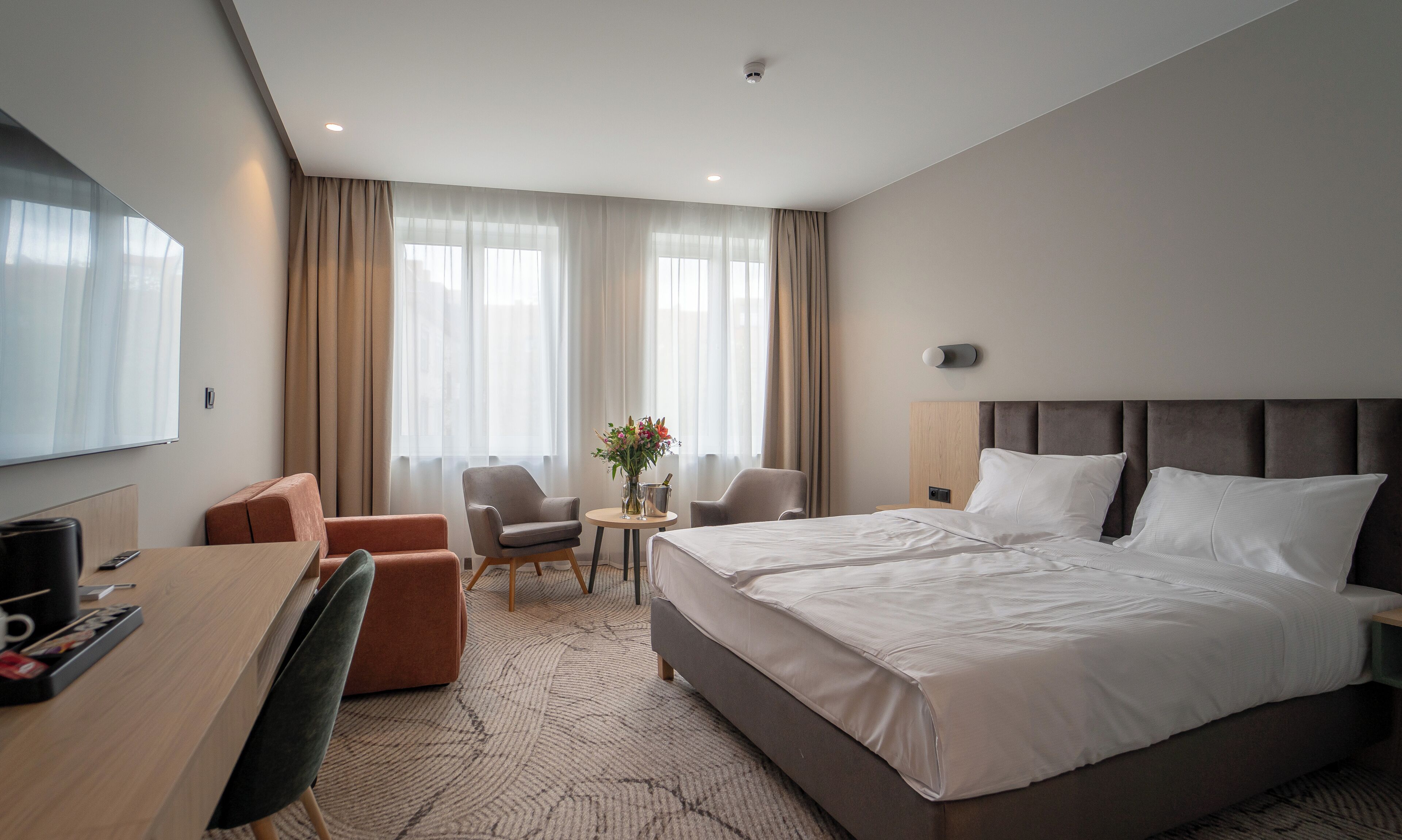 Habitación de hotel con cama, escritorio, silla, una mesita con un jarrón y ventana con cortinas.