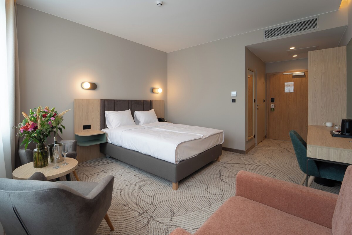 A hotel room with a bed, bedside tables, a desk, a chair, and a small table with a vase of flowers.