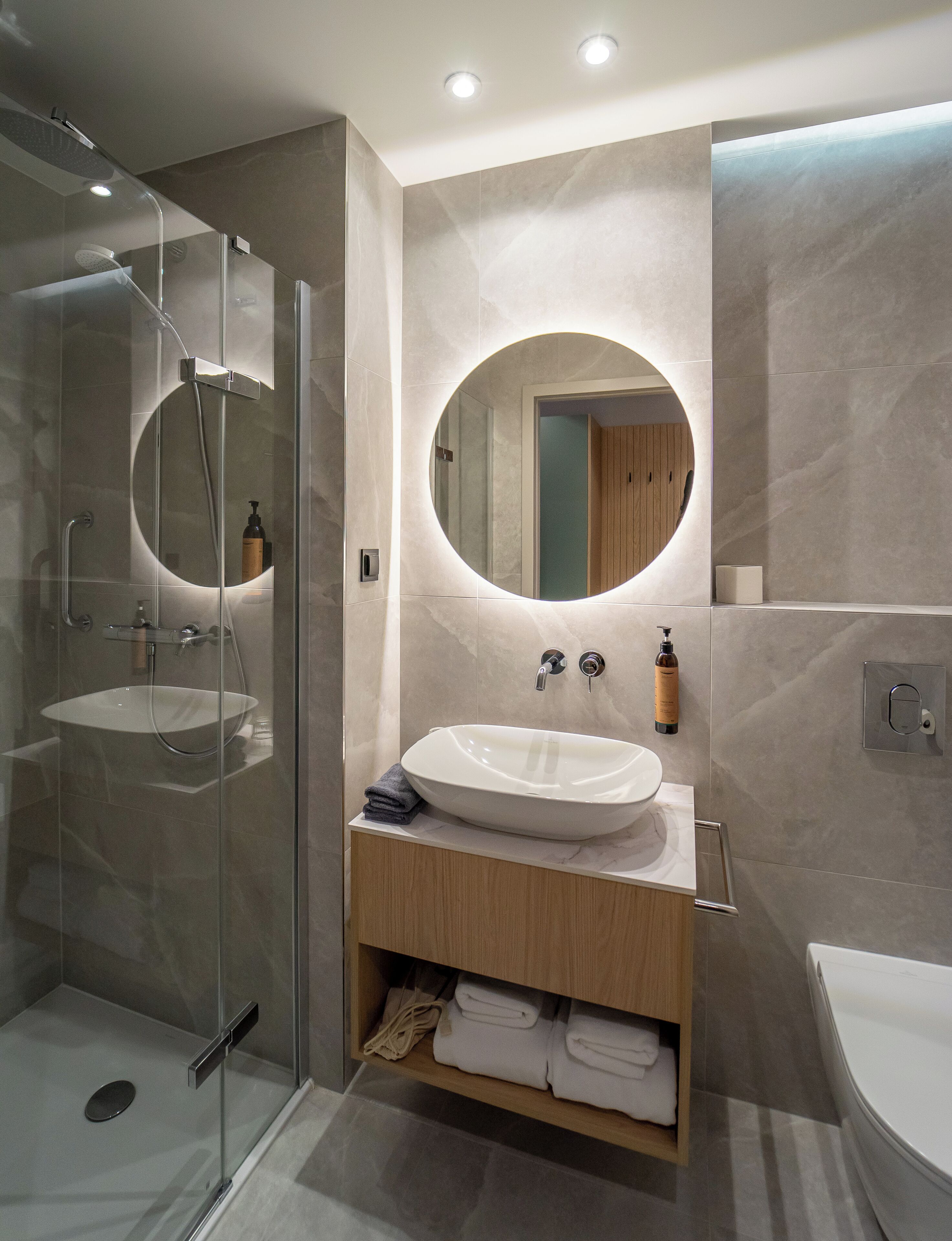 A modern bathroom with a glass-enclosed shower, a round mirror, and a white sink with a wooden vanity.