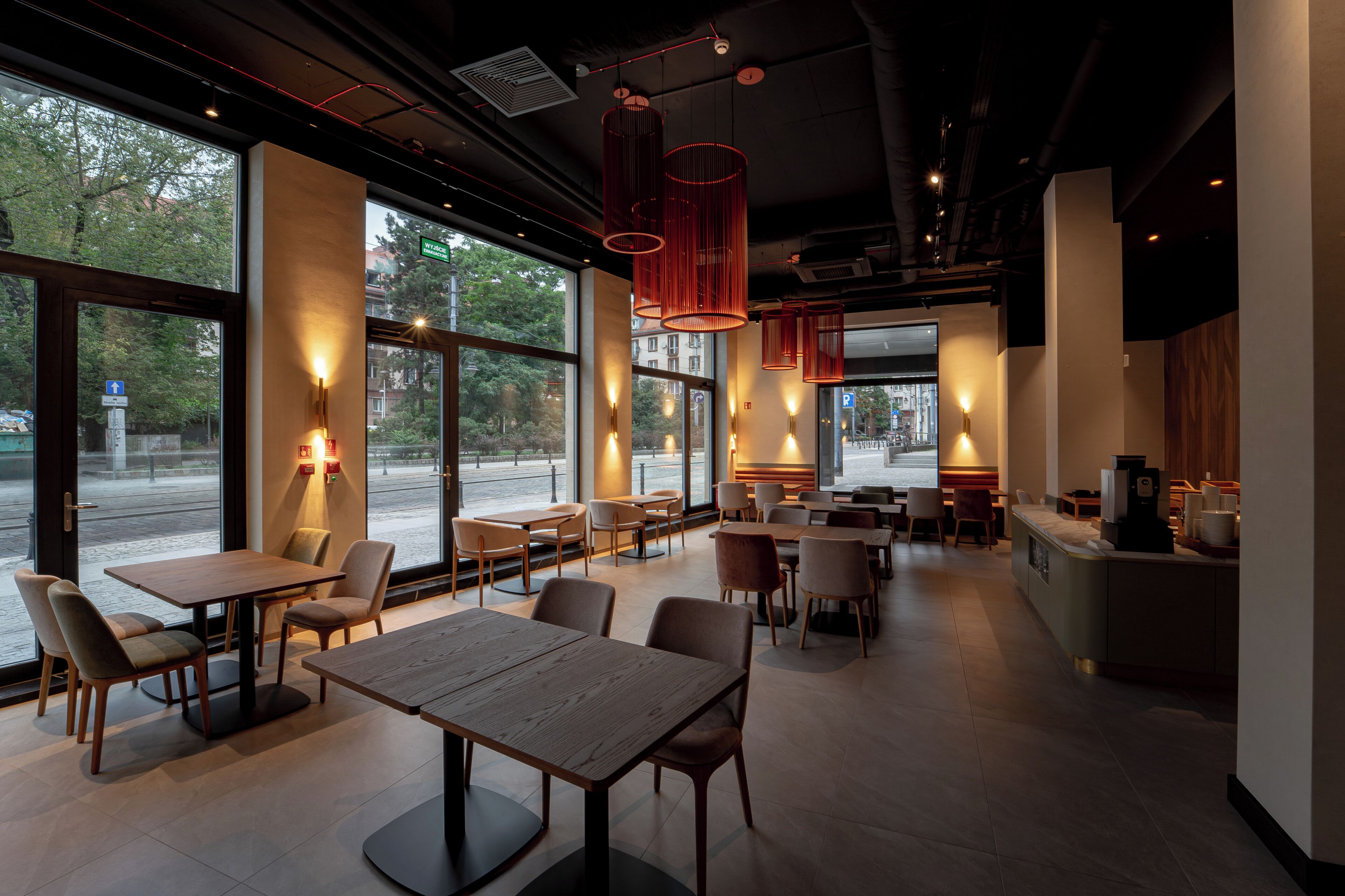 Un intérieur de restaurant moderne, avec de grandes fenêtres, des tables en bois et des luminaires suspendus rouges.