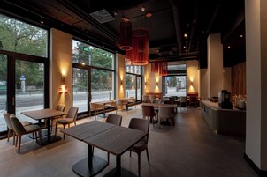 A modern restaurant interior with large windows, wooden tables, and red pendant lighting.
