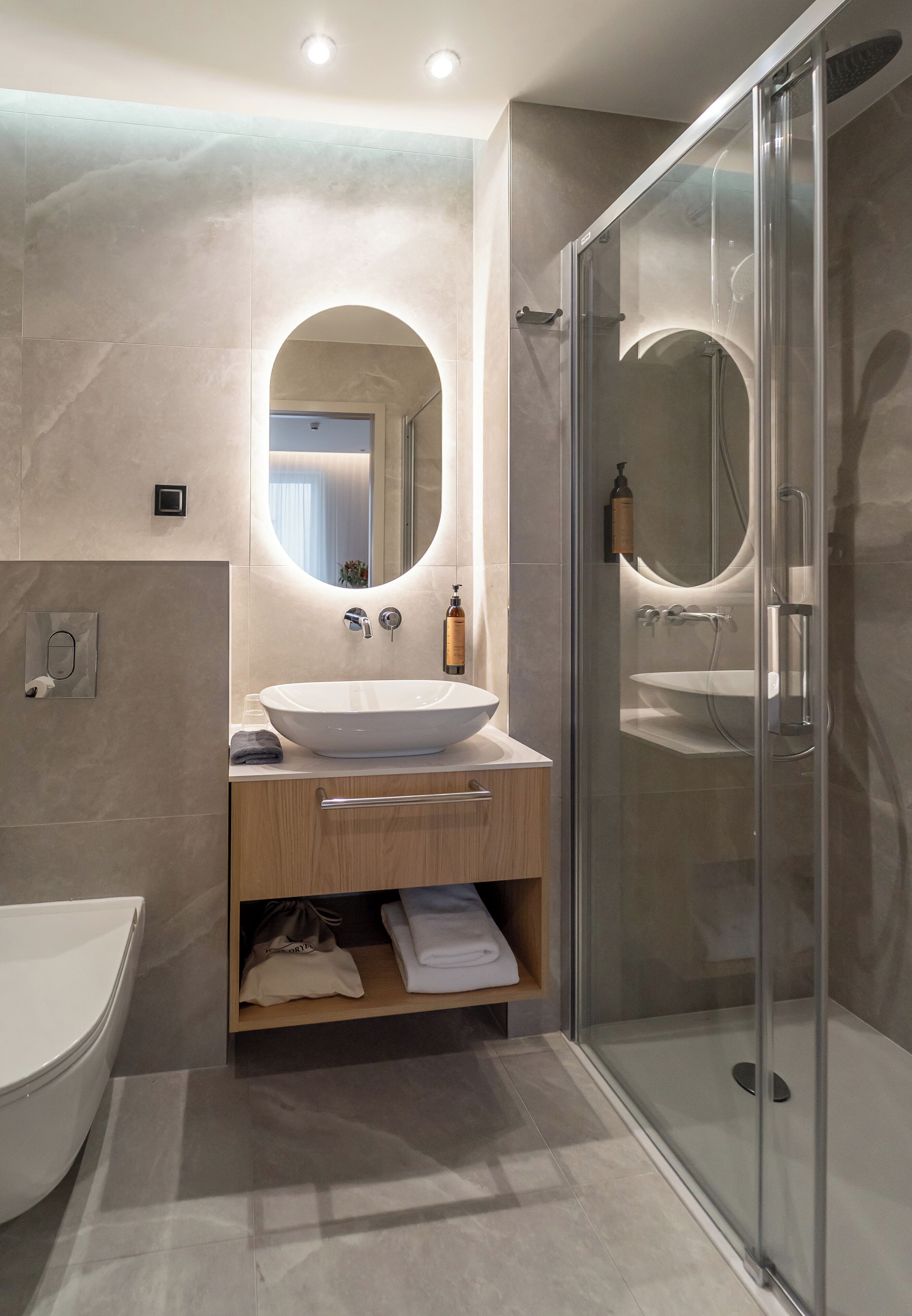 Ein modernes Badezimmer mit einem großen, ovalen Spiegel, einem weißen Waschbecken und einer Duschkabine aus Glas.