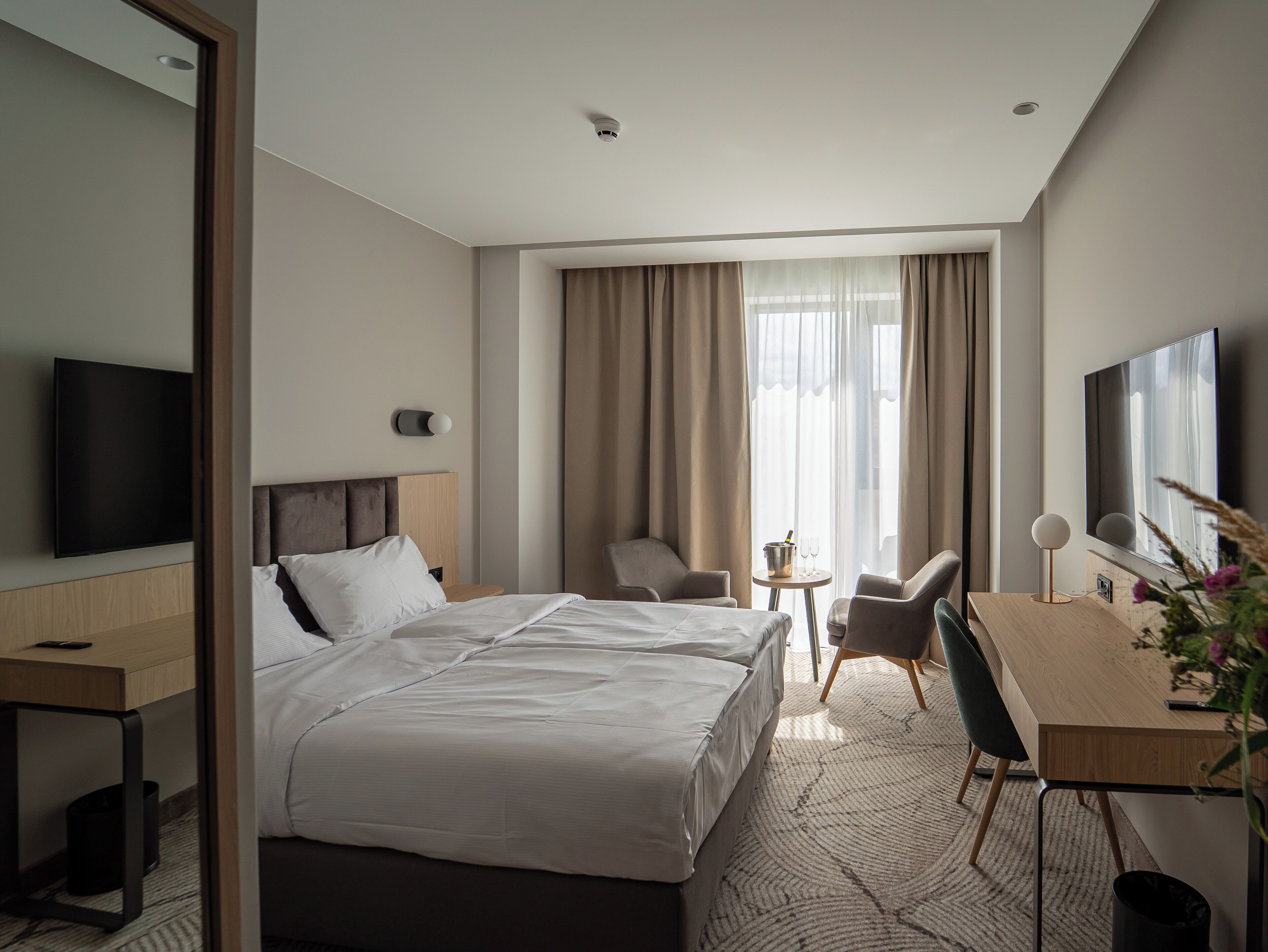 Quarto de hotel com cama, escrivaninha, cadeira, televisão e janela com cortinas.