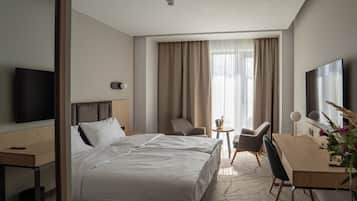 Quarto de hotel com cama, escrivaninha, cadeira, televisão e janela com cortinas.