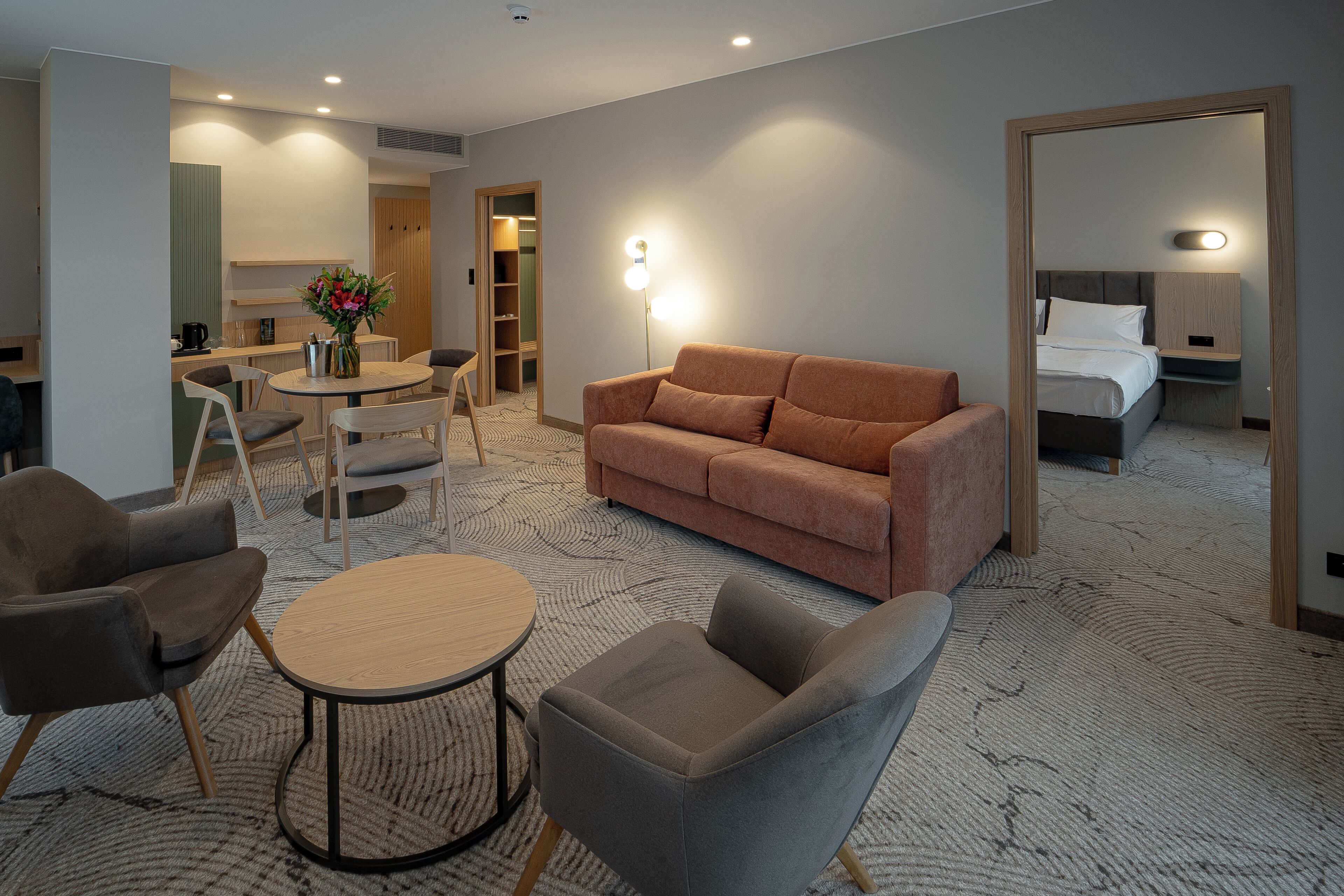 A hotel room with a sofa, armchairs, a small table, a bed, and a bathroom visible through an open door.