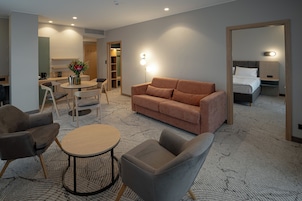 Habitación de hotel con sofá, sillones, una mesita, cama y baño visible a través de una puerta abierta.
