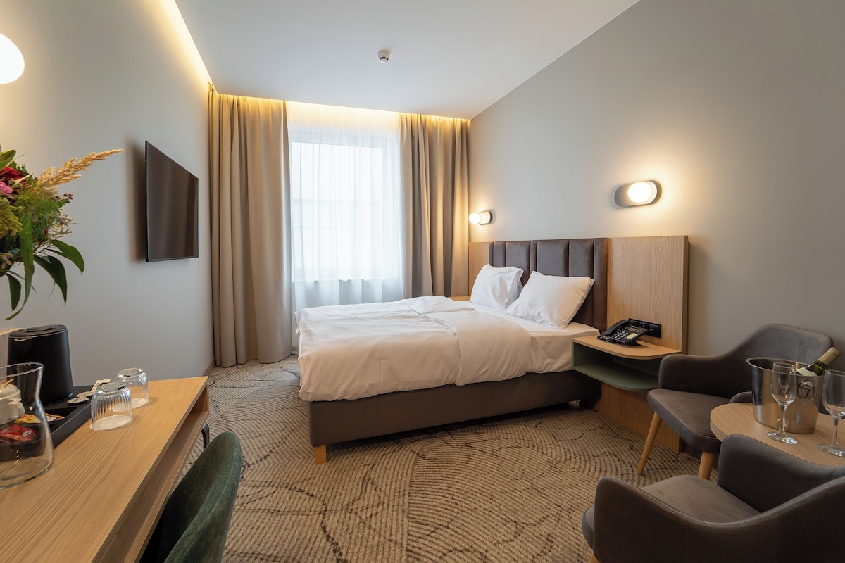Hotellihuone, jossa on sänky, yöpöytä, tuoli, työpöytä ja suuri ikkuna, jossa on verhot.