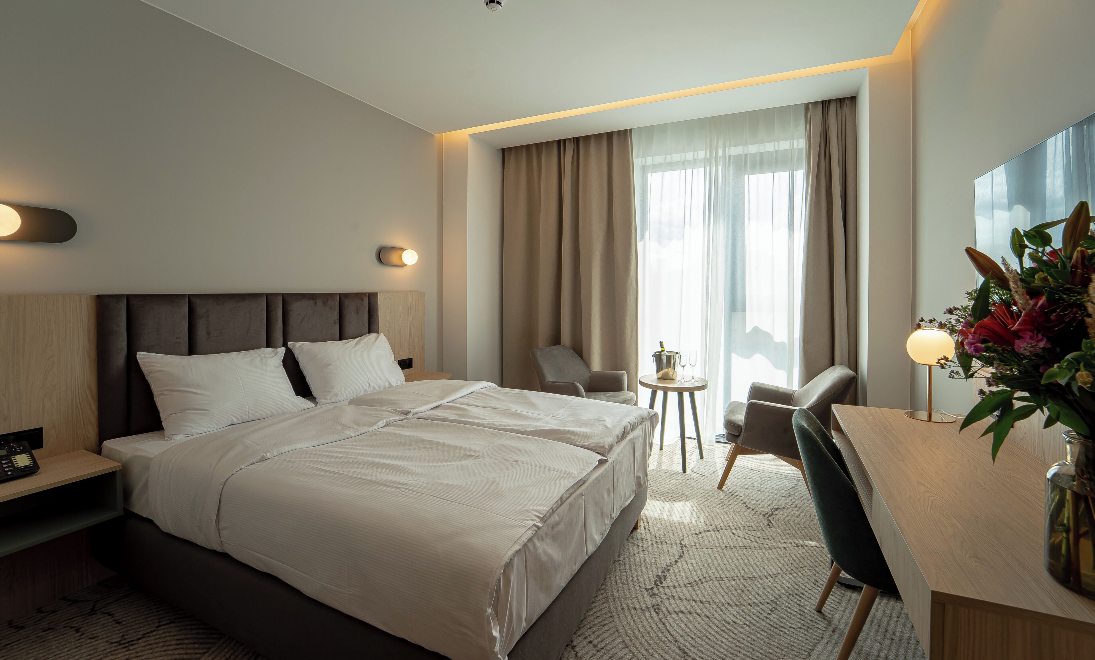 Une chambre d’hôtel avec un lit, deux chaises, un bureau, une petite table avec une bouteille et des verres, et une grande fenêtre avec des rideaux.