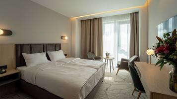 Une chambre d’hôtel avec un lit, deux chaises, un bureau, une petite table avec une bouteille et des verres, et une grande fenêtre avec des rideaux.