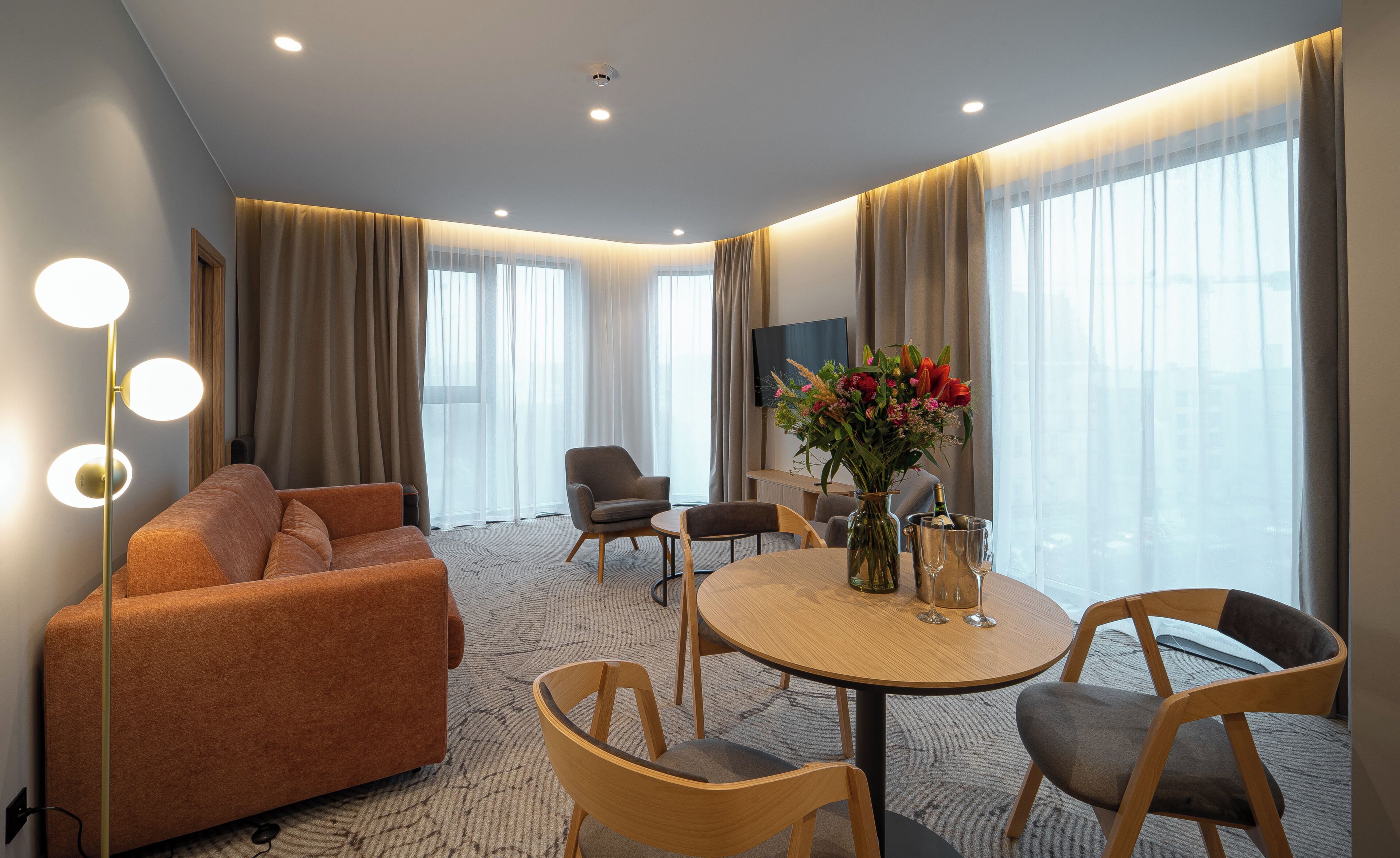 Une chambre d’hôtel moderne, équipée d’un canapé, de fauteuils, d’une table à manger et d’une grande fenêtre avec des rideaux.