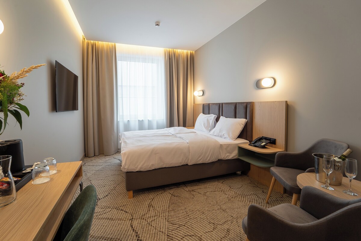 A hotel room with a bed, bedside table, chair, desk, and a large window with curtains.