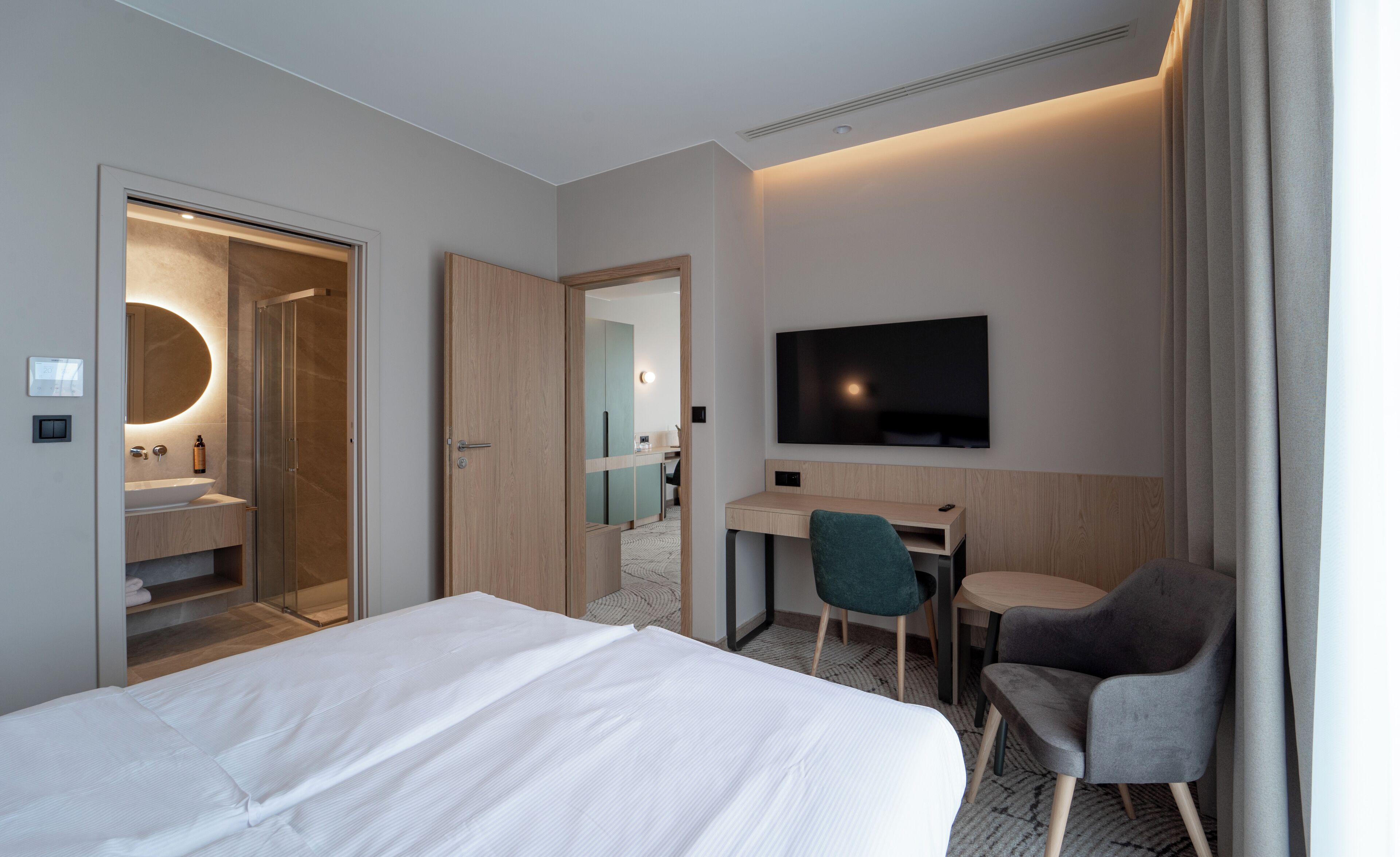 A hotel room with a bed, a desk, a chair, a TV, and a bathroom with a mirror and sink.