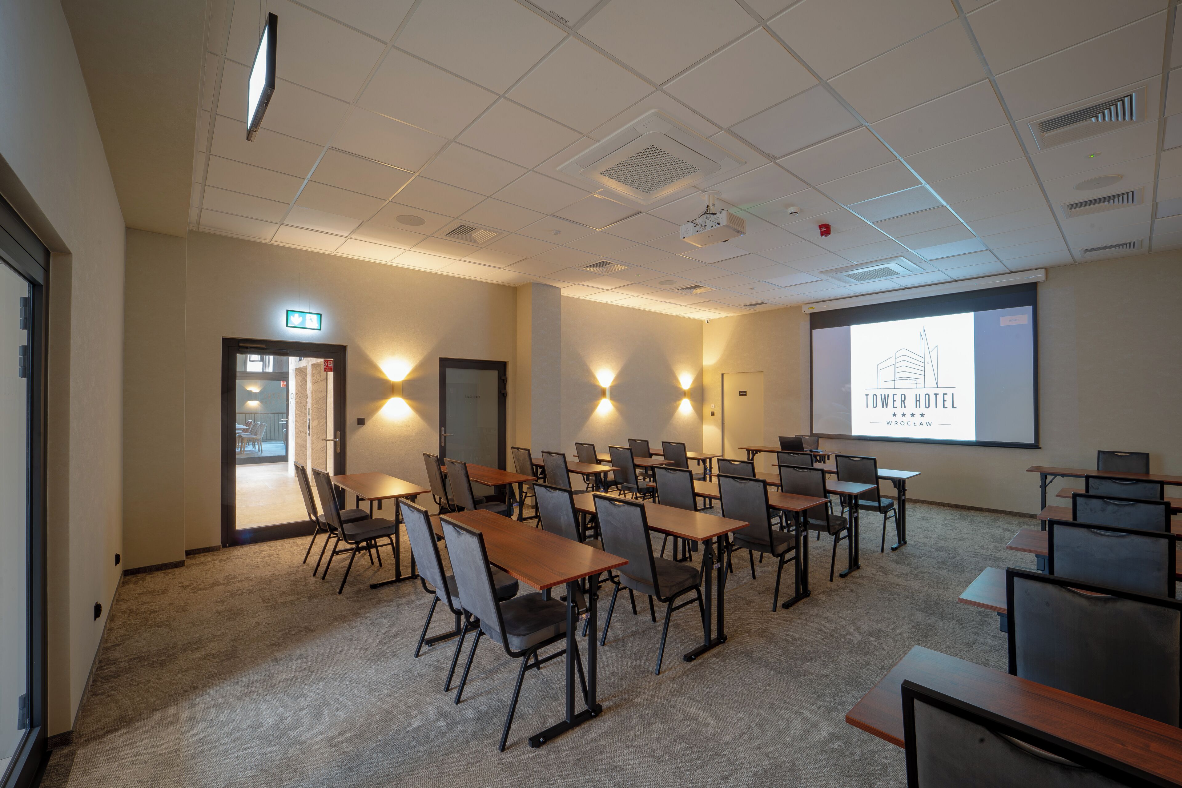 Un salón de conferencias con mesas y sillas, una pantalla de proyector y una puerta con letrero de salida.