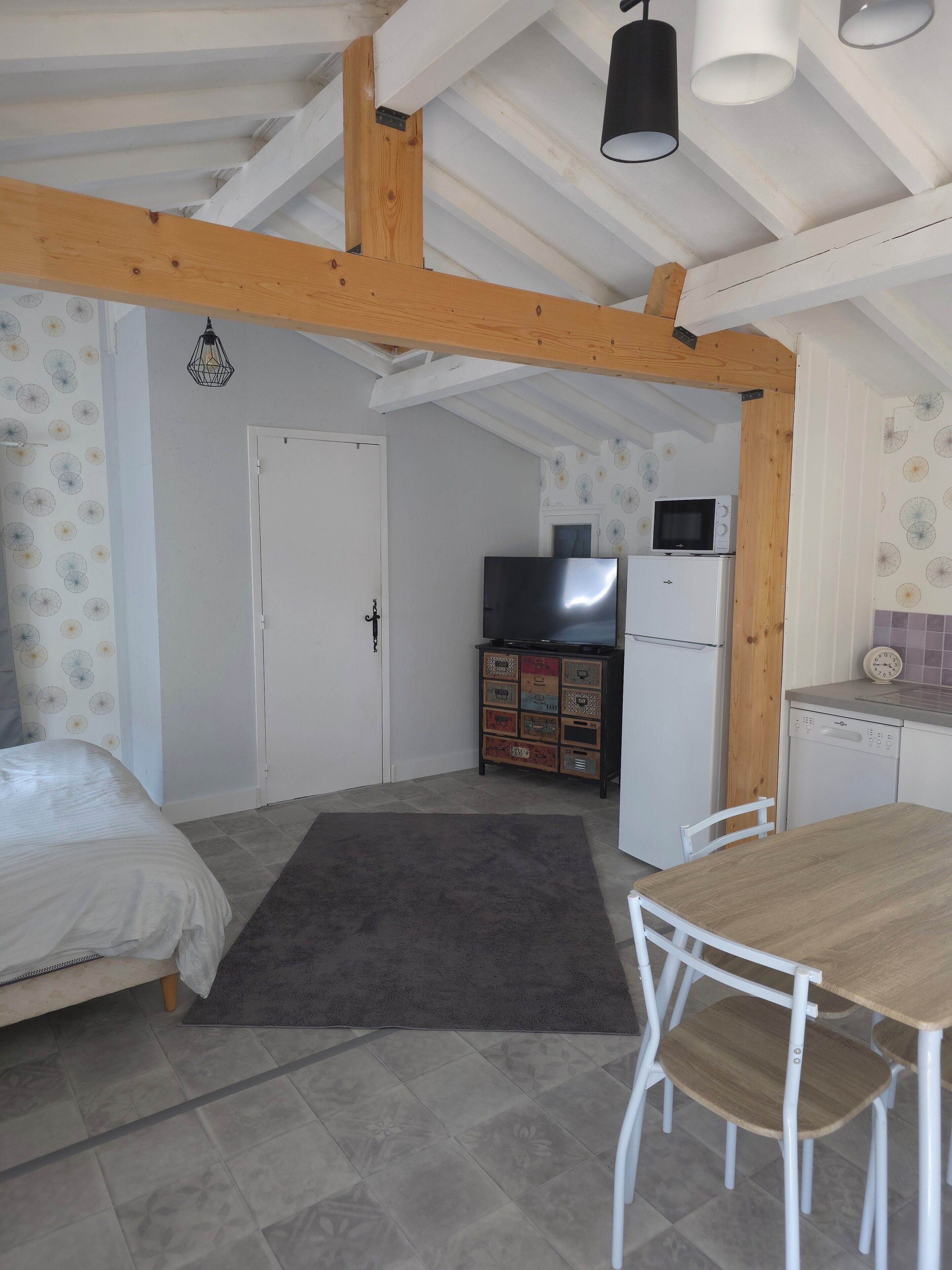 A room with a bed, a table, chairs, a kitchen area with appliances, and a ceiling with exposed beams.