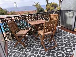 Een balkon met twee houten stoelen en een tafel, met uitzicht op een zwembad en de zee.
