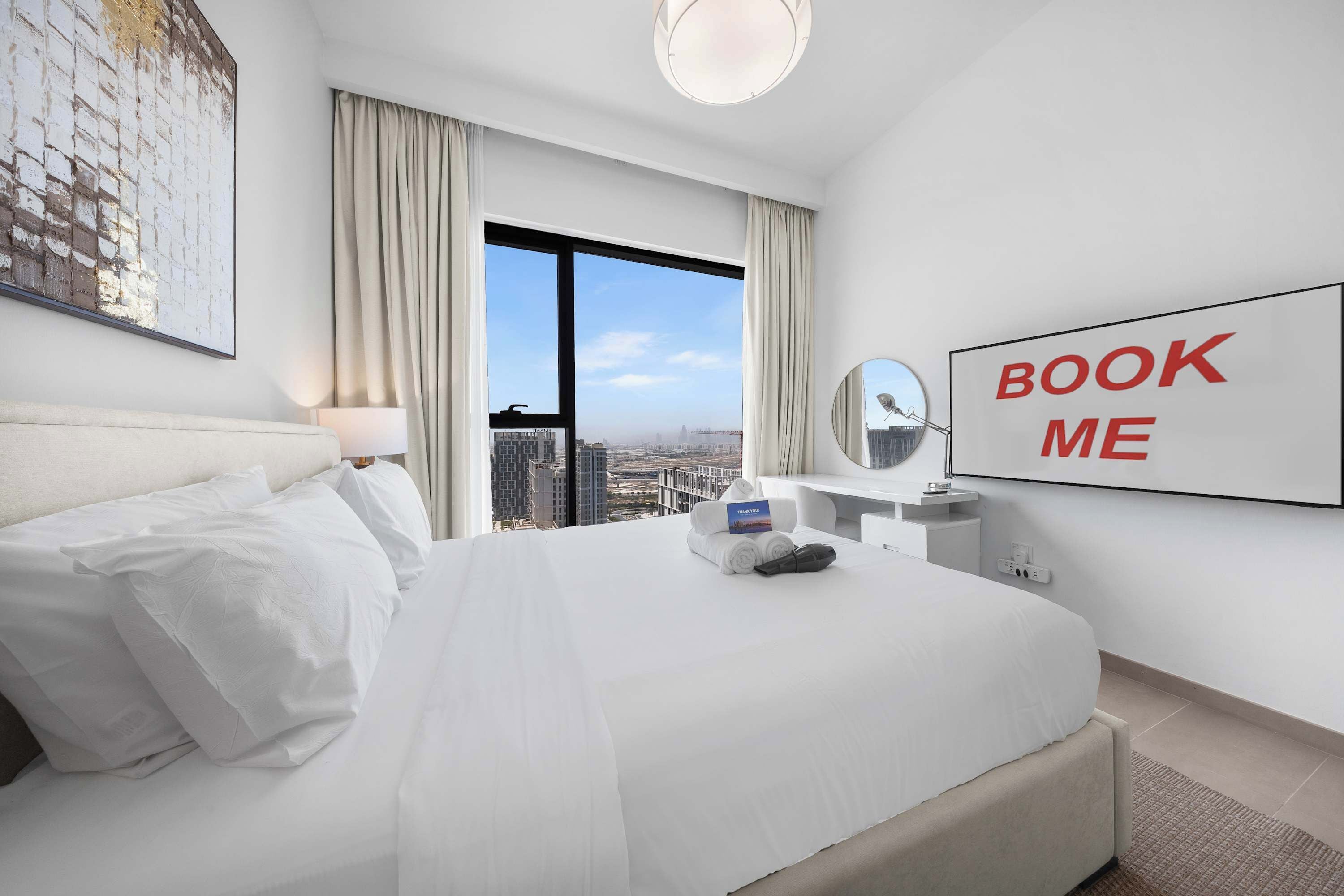 Hotelkamer met een groot bed, een bureau met spiegel, een bordje met de tekst 'BOOK ME' en uitzicht op de stad.