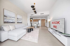 A modern living room with a white sofa, a coffee table, and a TV on a white stand. There are two framed pictures on the wall, a chandelier, and a kitchen in the background. - StayShort - Chic 1-Bedroom for 4 in Prime Dubai Hills Location! (Dubai Hills)