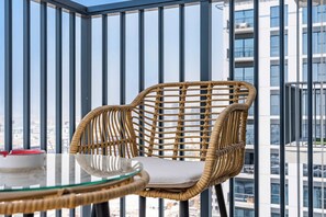 A wicker chair with a cushion on a balcony with a glass table, overlooking a cityscape. - StayShort - Chic 1-Bedroom for 4 in Prime Dubai Hills Location! (Dubai Hills)