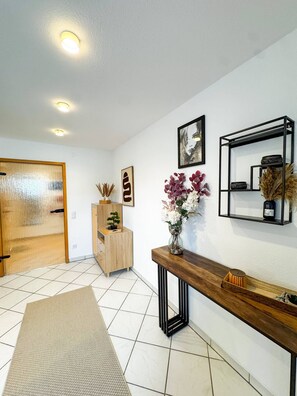 A modern interior with a wooden door, a small wooden cabinet, a console table with a vase of flowers, a framed picture, and a black metal shelf with decorative items. - Bibo City Suite Apartment (Bielefeld)