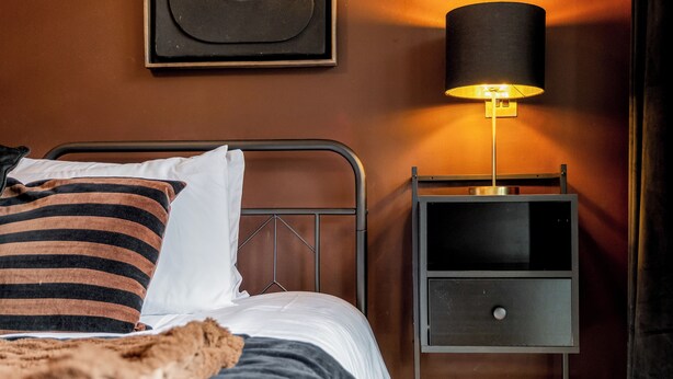 Ein ordentlich bezogenes Bett mit gestreiftem Kissen, eine Nachttischlampe und ein kleiner Nachttisch mit Schublade.