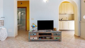 Un soggiorno con televisione su un mobile in legno, una cucina con mobili bianchi e un bagno con doccia in vetro.