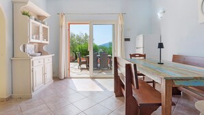 Zona pranzo con tavolo e sedie, cucina con mobili e vista su un patio esterno attraverso una porta vetrata.