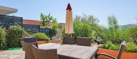 An outdoor patio with a table and chairs, surrounded by greenery and a clear sky.