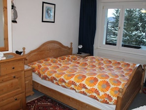 A bedroom with a wooden bed, a dresser, a nightstand, a framed picture on the wall, and a window with a view of snow-covered trees. - Haus Seewaldweg 309 Appartement/Fewo,  , 3-Bettwohnung 3-Bettwohnung by Interhome (Arosa)