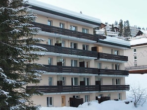 Um hotel com vários andares, varandas, telhado coberto de neve e um pinheiro em primeiro plano.