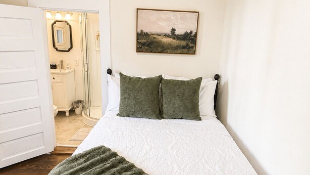 Une chambre à coucher avec un lit, un plaid vert, un tableau accroché au mur et une salle de bain visible à travers une porte ouverte.