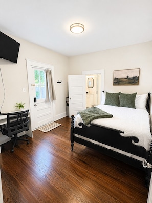 A bedroom with a bed, a desk, and a chair. There is a door leading to a bathroom, and a painting on the wall.