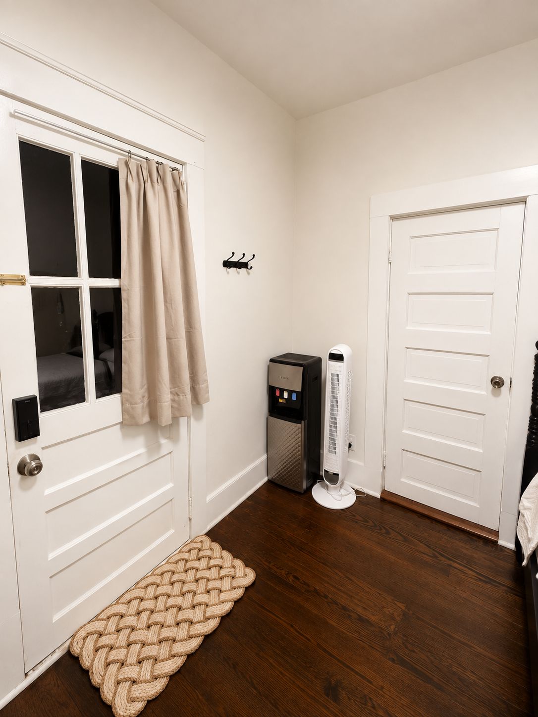 Une pièce avec une porte blanche, une fenêtre ornée de rideaux beiges, un petit radiateur et un tapis tissé sur le sol.