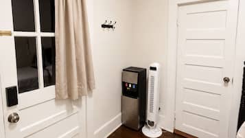 Une pièce avec une porte blanche, une fenêtre ornée de rideaux beiges, un petit radiateur et un tapis tissé sur le sol.