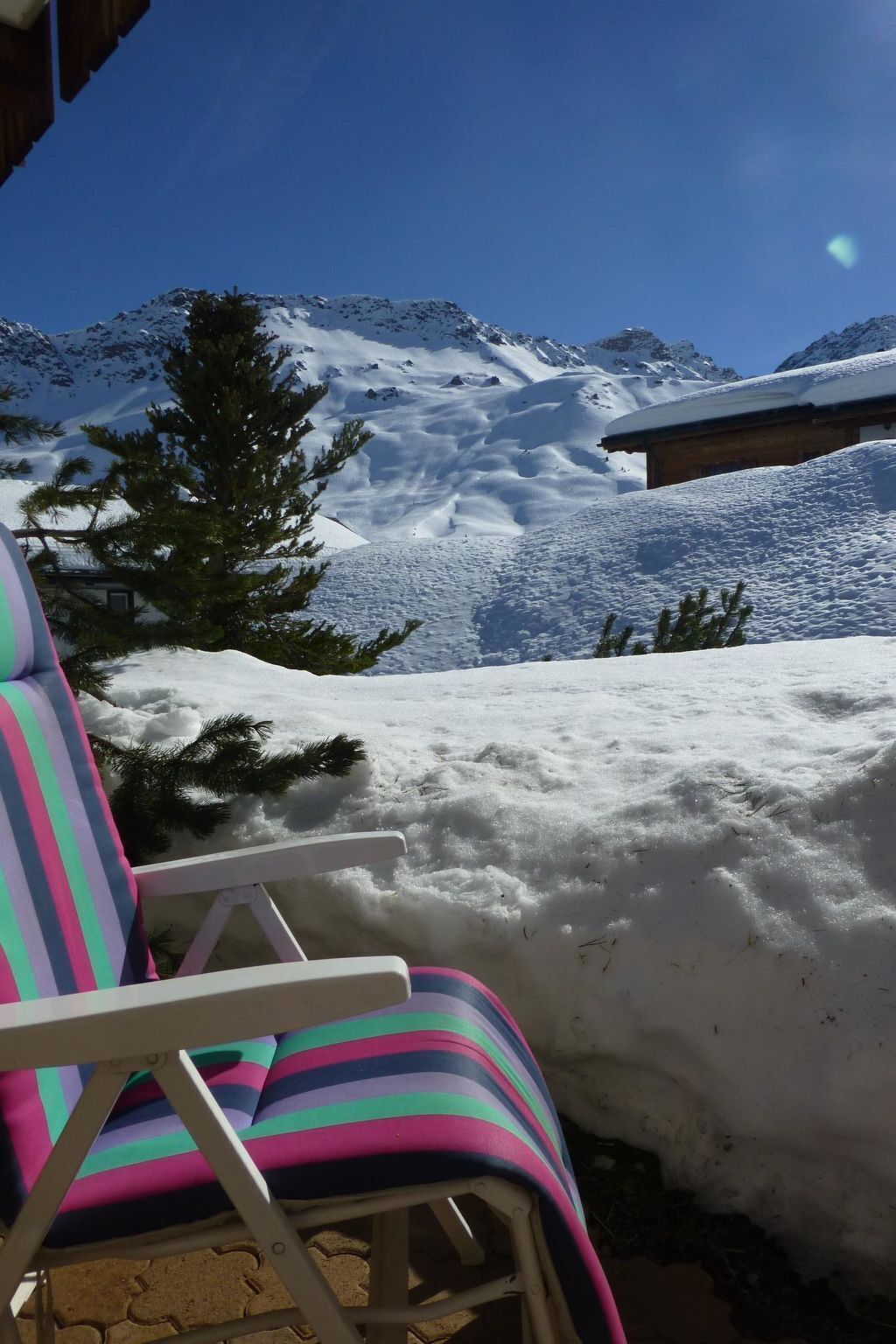 Uma cadeira com almofada listrada e colorida, uma montanha coberta de neve ao fundo e um céu azul claro.