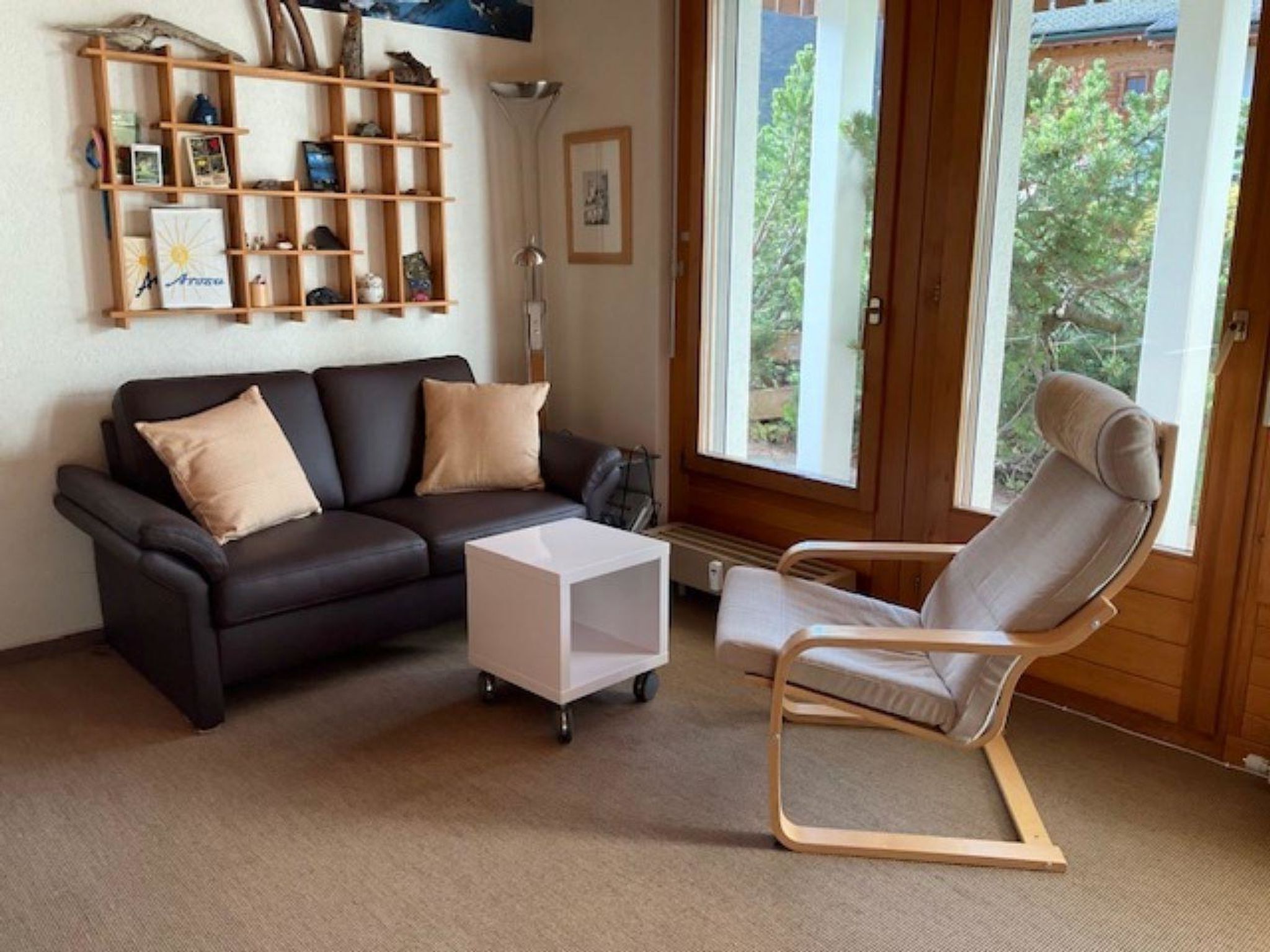 A living room with a dark leather sofa, a white side table, a wooden bookshelf, and a window with a view of greenery.