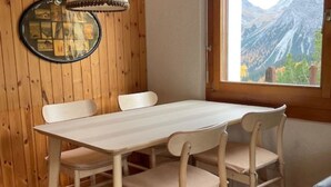 Ett rum med träpanel på väggarna, ett bord och stolar, en hängande lampa, en inramad tavla och ett fönster med utsikt över bergen.