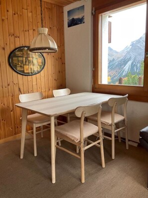 Ein Zimmer mit Holzwänden, einem Tisch, Stühlen, einer Hängelampe, einem gerahmten Bild und einem Fenster mit Blick auf die Berge.