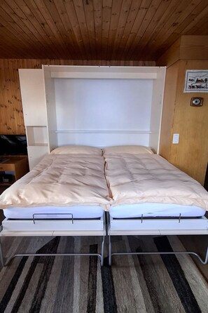 Ein Bett mit weißem Kopfteil und Metallgestell, ein Streifenmuster-Teppich auf dem Boden und eine Holzbalkendecke.