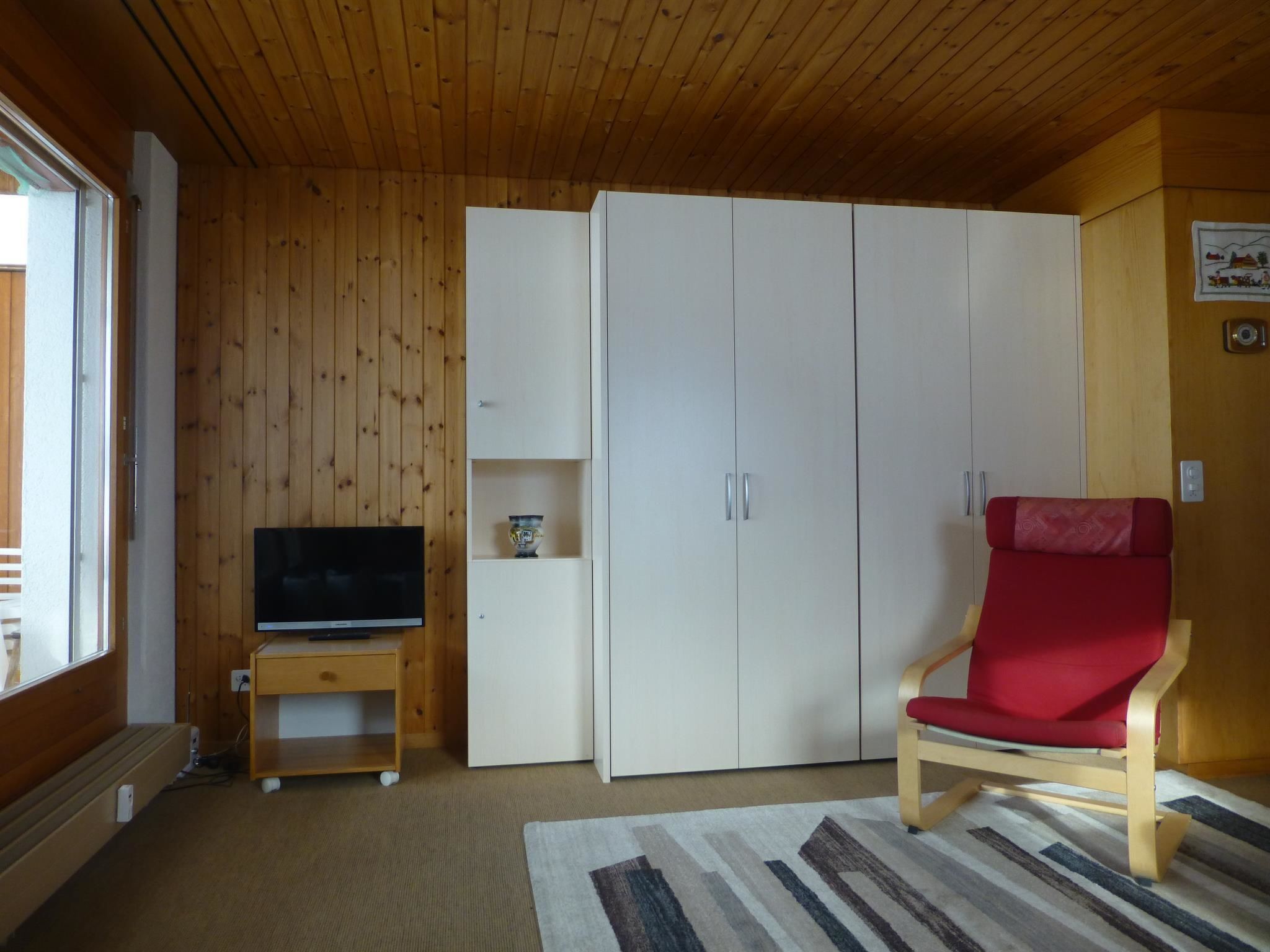 A room with a television on a wooden stand, a wardrobe, a red chair, and a patterned rug.