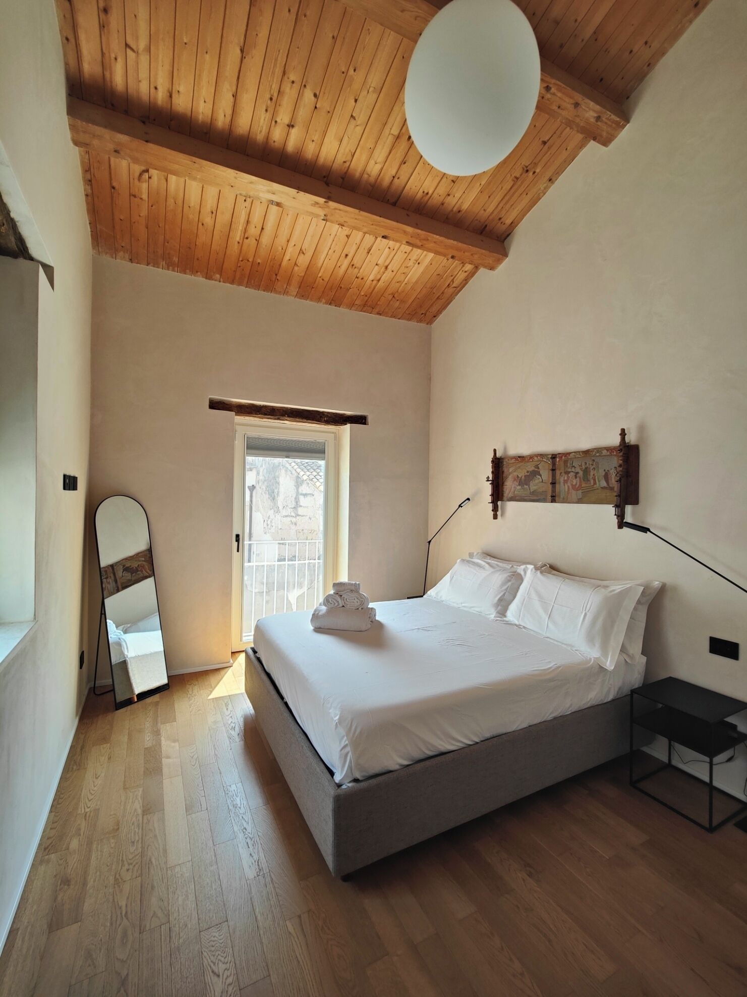 A bedroom with a bed, a bedside table, a mirror, and a door leading to an outdoor area.