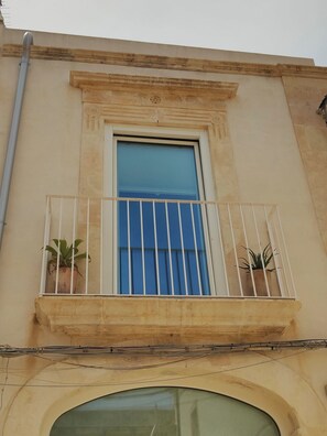 Ein Gebäude mit Balkon, das ein Fenster mit vertikalen Gitterstäben, zwei Blumentöpfe und eine sichtbare Leitungsrohr zeigt.