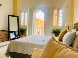Une chambre avec un lit, une commode, un miroir, deux plantes en pot et une vue sur le balcon.
