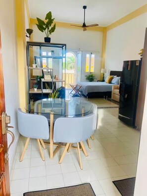 Un coin repas avec une table en verre et quatre chaises, un canapé-lit à l’arrière-plan et un réfrigérateur sur la droite.