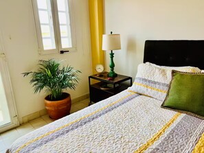Un lit bien fait, recouvert d’une couvre-lit à rayures, avec un oreiller vert et une table de chevet sur laquelle se trouvent une lampe et une horloge.