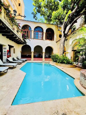 Ein Schwimmbecken mit Fliesen umgeben, umrahmt von einem Gebäude mit Rundbogenfenstern und einem Balkon mit Blumen.