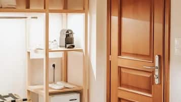 A room with a wooden door, a small white refrigerator, and a wooden shelf unit with a kettle and cups.