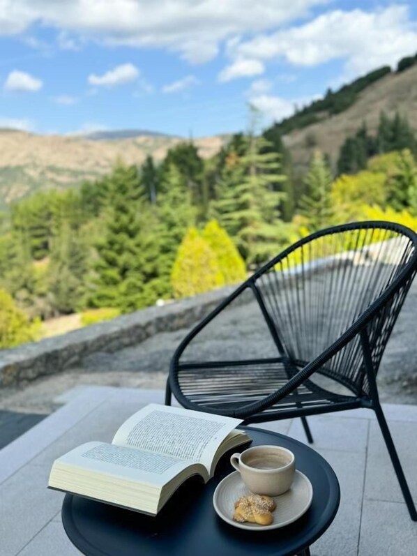 An open book on a table with a cup and a cookie, set outdoors with a chair and a view of trees and hills.